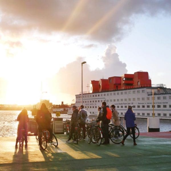 A group of people on an Amsterdam bike tour stand with their bicycles on a waterfront platform at sunset, with a large ship docked in the background.