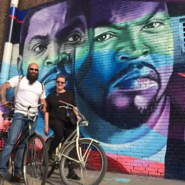 Two people with bicycles stand in front of a colorful mural on a brick wall, featuring two large, realistic faces—capturing the vibrant energy of an Amsterdam urban bike tour.