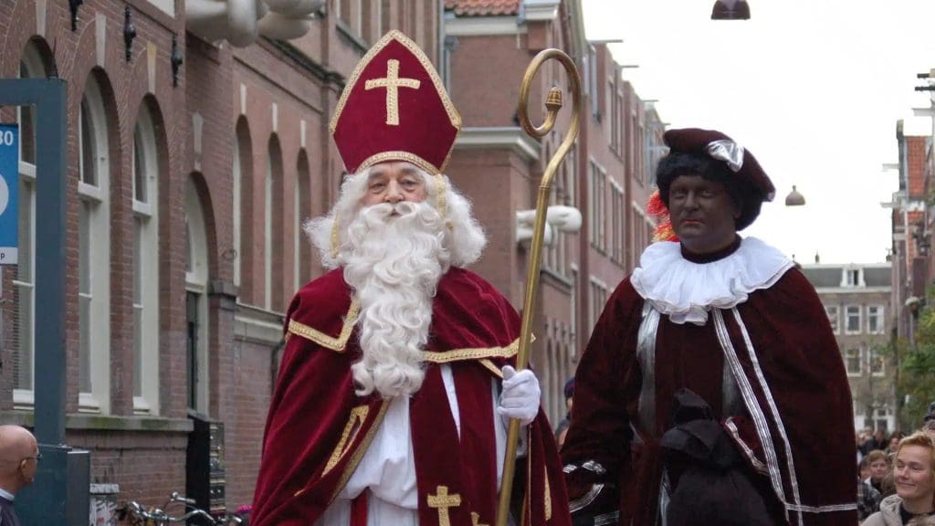 Two people in costume walk down a street: one dressed as Sinterklaas in red robes and miter, the other as Zwarte Piet in dark makeup and traditional attire.
