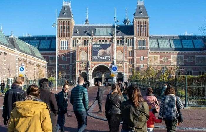 A group of people walk toward the main entrance of the Rijksmuseum, a large historic brick building in Amsterdam, on a clear day.