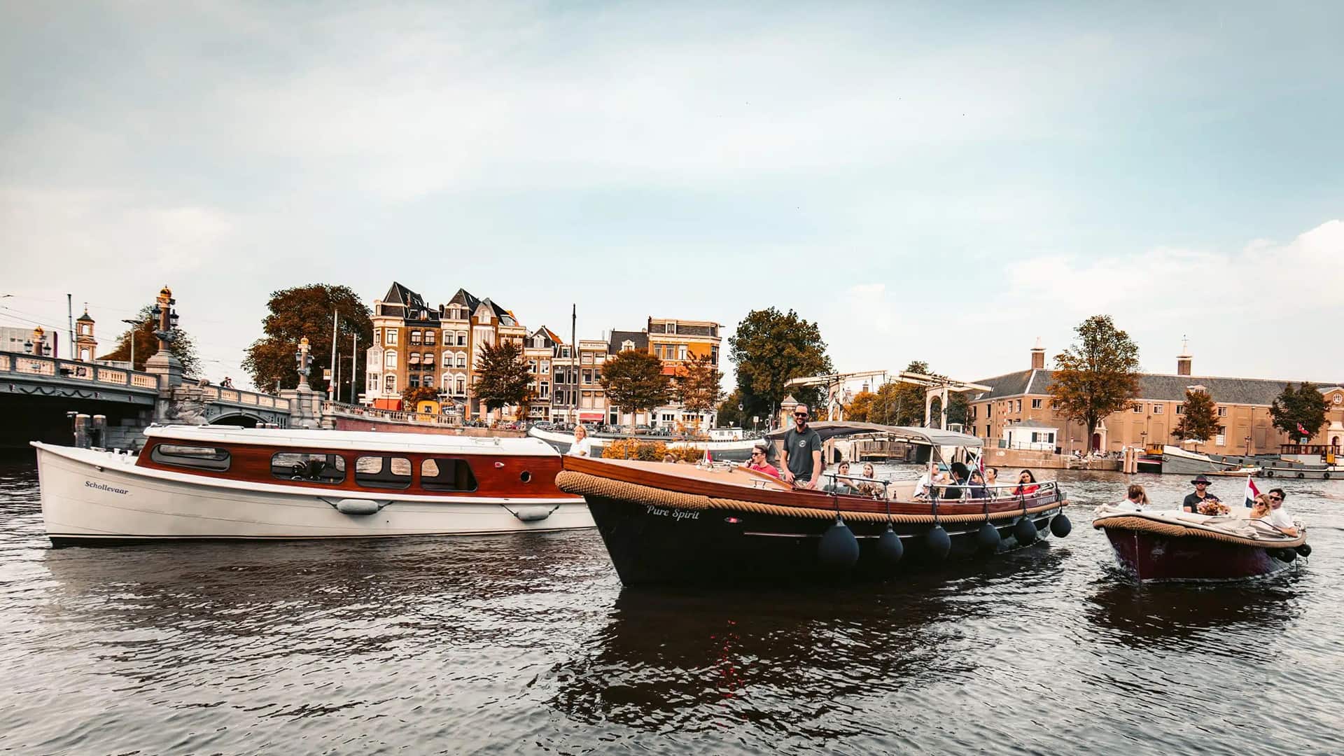 Three boats with people onboard are floating on a calm river near a city shoreline with trees and historic buildings in the background.