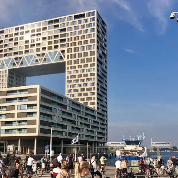 Modern apartment building with a large rectangular gap above a busy waterfront area, where people walk and ride bikes, with boats visible in the background under a clear sky.
