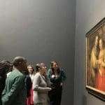 A group of people observe a framed painting displayed on a museum wall; some read the nearby information plaque.