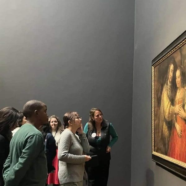 A group of people observe a framed painting displayed on a museum wall; some read the nearby information plaque.