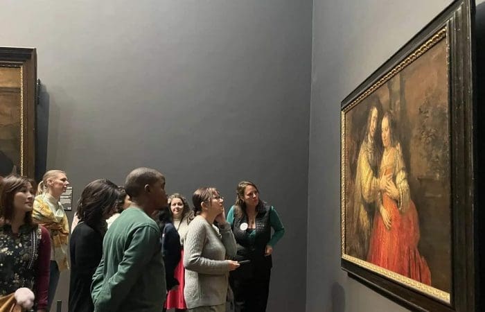 A group of people observe a framed painting displayed on a museum wall; some read the nearby information plaque.