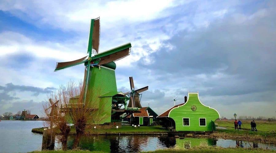 Green windmills and buildings sit beside a canal under a partly cloudy sky, with several people walking along the water’s edge. Green windmills and buildings sit beside a canal under a partly cloudy sky, with several people walking along the water’s edge.