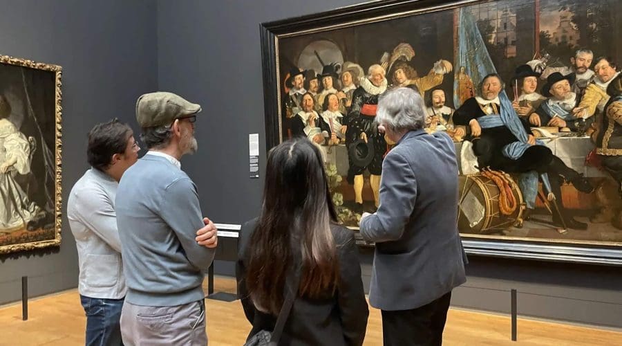 Four people stand in front of a large framed painting in a museum, listening to a guide explain the artwork. Four people stand in front of a large framed painting in a museum, listening to a guide explain the artwork.