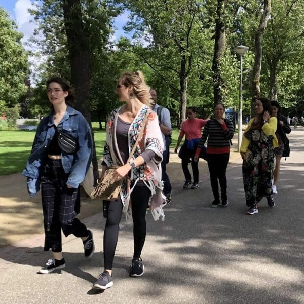A group of people walk along a paved path in a park with trees and grass on a sunny day.