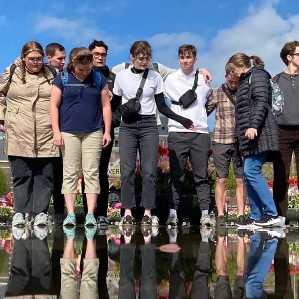 A group of people stands closely together by a reflective pool outdoors, with flowers and buildings in the background under a partly cloudy sky.