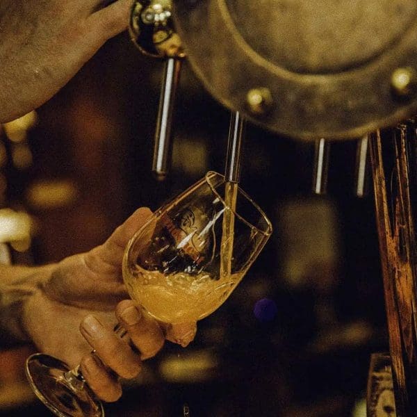 A person holds a glass at an angle while pouring beer from a tap in a dimly lit bar or brewery setting.