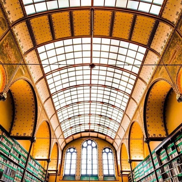 Library interior with tall bookshelves on both sides, arched ceiling with glass panels, and large windows at the far end letting in natural light.