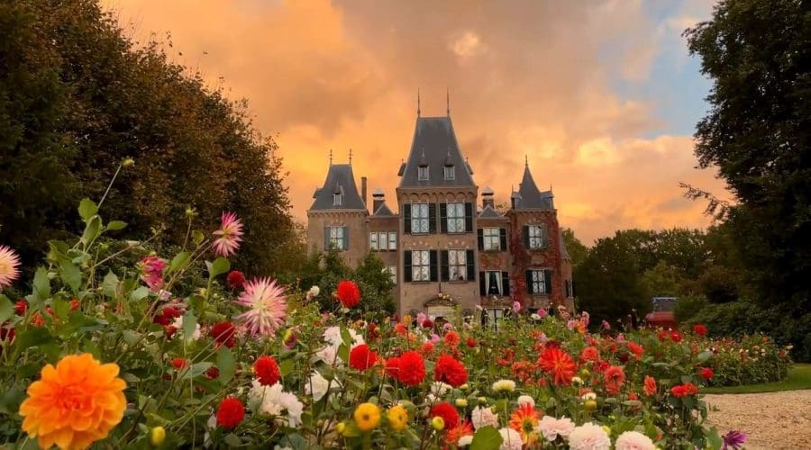A large stone building with pointed roofs stands behind a vibrant garden filled with colorful flowers under an orange sky at sunset.