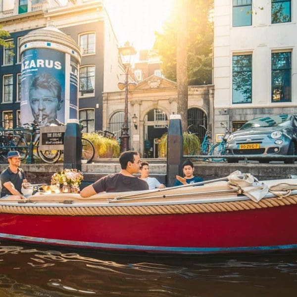 A red boat with four people cruises along an Amsterdam canal, passing parked cars and buildings, with sunlight shining in the background.