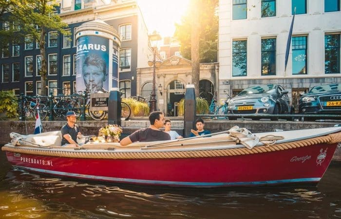 A red boat with four people cruises along an Amsterdam canal, passing parked cars and buildings, with sunlight shining in the background.