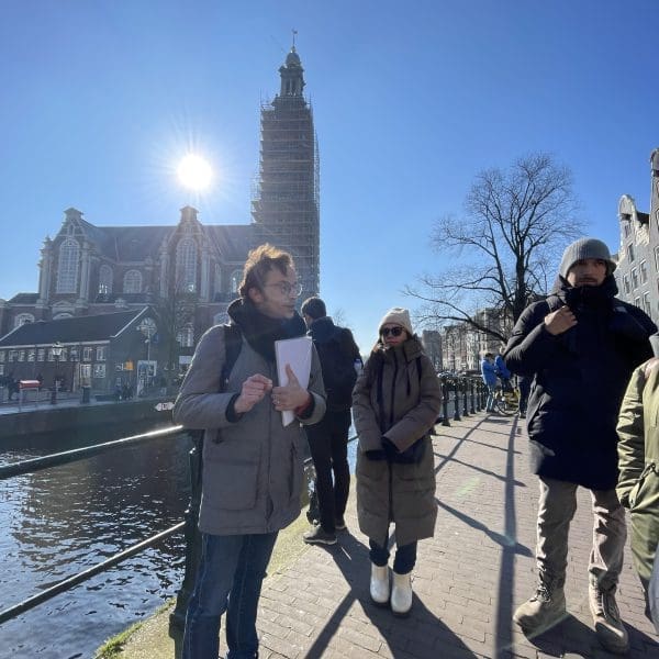 A group of people stand on a canal-side path in a city with a church and a scaffolded tower in the background under a clear blue sky.