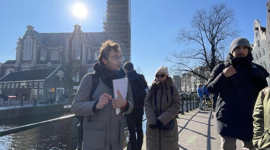 A group of people stand on a canal-side path in a city with a church and a scaffolded tower in the background under a clear blue sky.