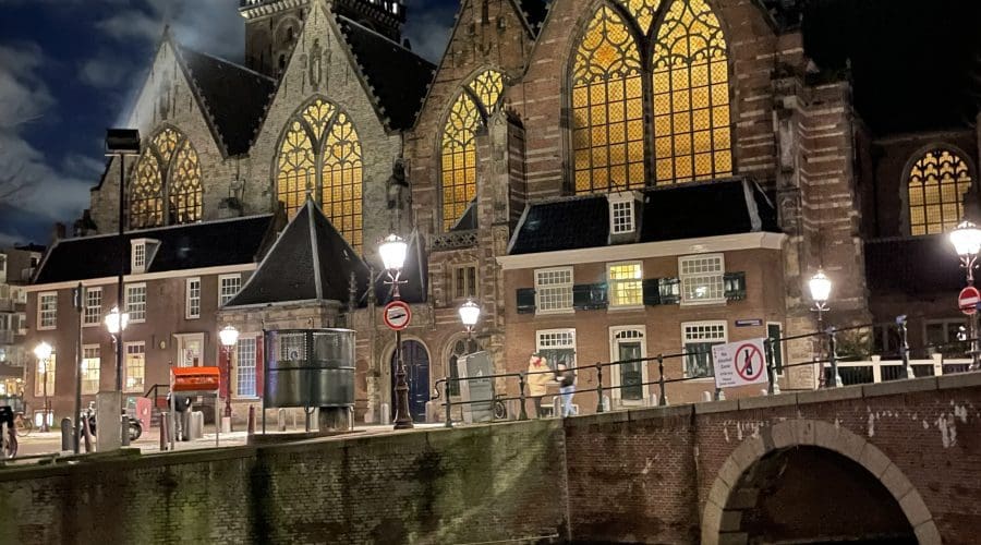 Night view of a historic church with illuminated stained glass windows, pointed arches, and a tall clock tower, reflected in a canal below. Night view of a historic church with illuminated stained glass windows, pointed arches, and a tall clock tower, reflected in a canal below.