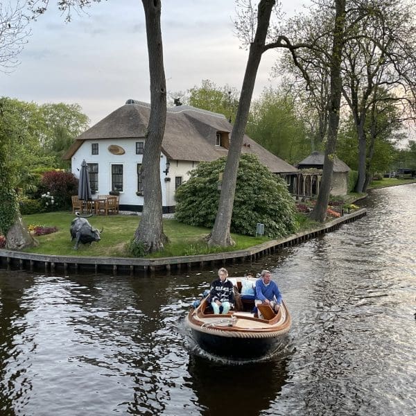 Two people ride a small motorboat along a canal bordered by trees, with a thatched-roof house and outdoor seating area visible in the background.