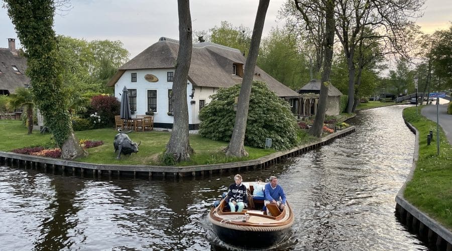 Two people ride a small motorboat along a canal bordered by trees, with a thatched-roof house and outdoor seating area visible in the background.
