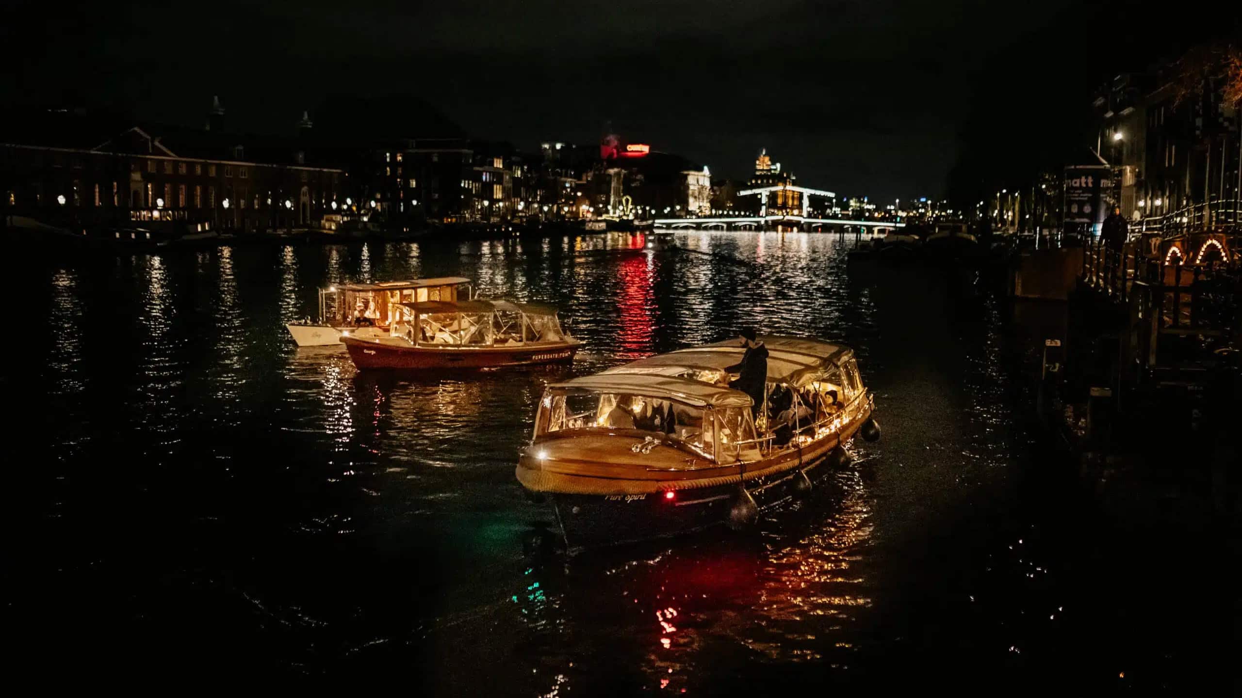 Two boats with transparent covers are floating on a calm, dark river at night, with city lights reflecting on the water in the background.
