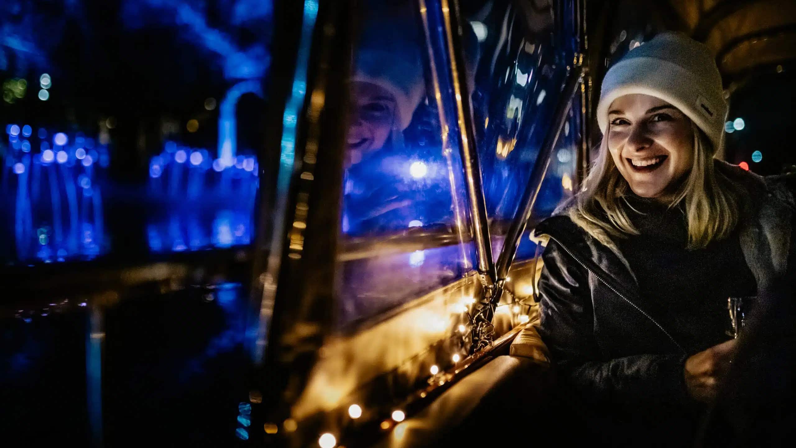 A woman wearing a beanie and coat sits beside a window at night, smiling with blue lights and her reflection visible in the glass.