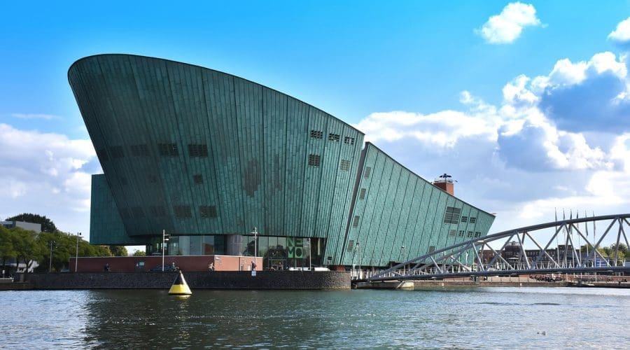 A modern green building with a slanted roof sits by the water, with a pedestrian bridge connecting it to the shore under a partly cloudy sky. A modern green building with a slanted roof sits by the water, with a pedestrian bridge connecting it to the shore under a partly cloudy sky.