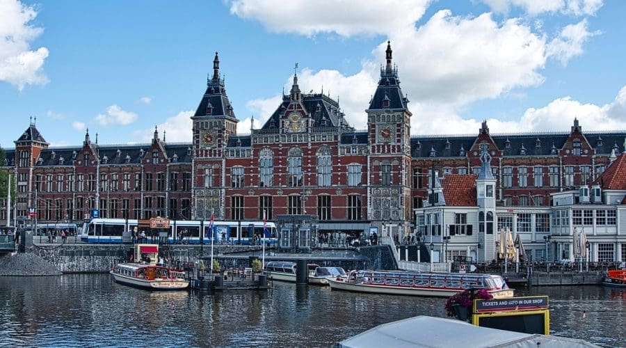 Amsterdam Central Station, a historic red-brick building with ornate towers, is seen across a canal with boats and trams in the foreground under a partly cloudy sky.