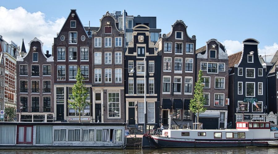 Row of traditional Dutch canal houses with gabled roofs and large windows, situated along a canal with houseboats moored in front. Row of traditional Dutch canal houses with gabled roofs and large windows, situated along a canal with houseboats moored in front.