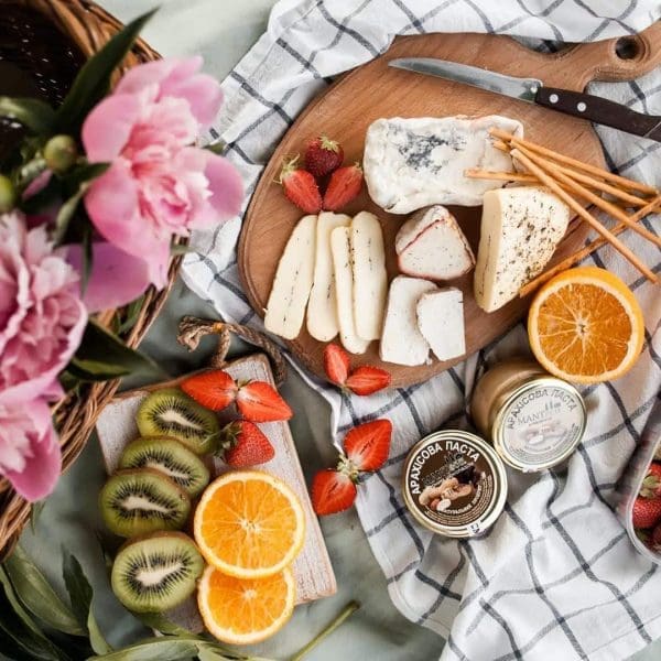 A picnic setup with various cheeses, strawberries, kiwi, orange slices, breadsticks, a knife, and pink flowers on a checkered cloth.