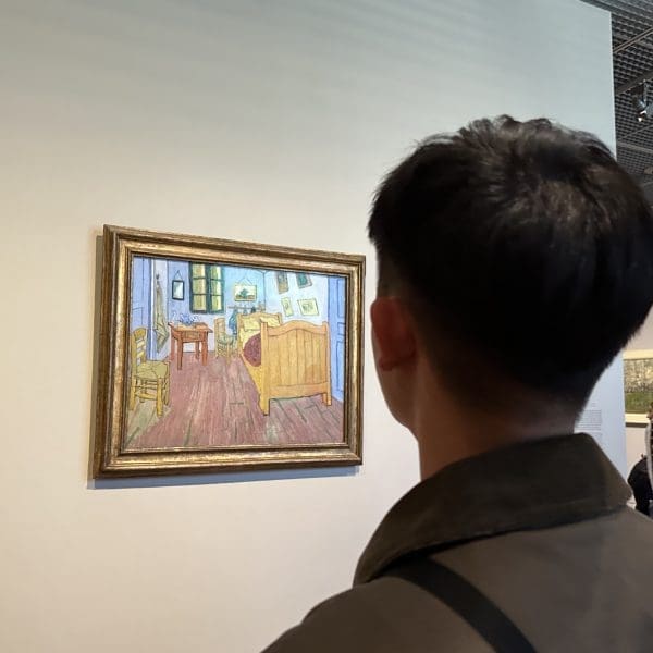 A person views Vincent van Gogh's painting "The Bedroom" in a museum, while another person takes a photo of the artwork with a smartphone.