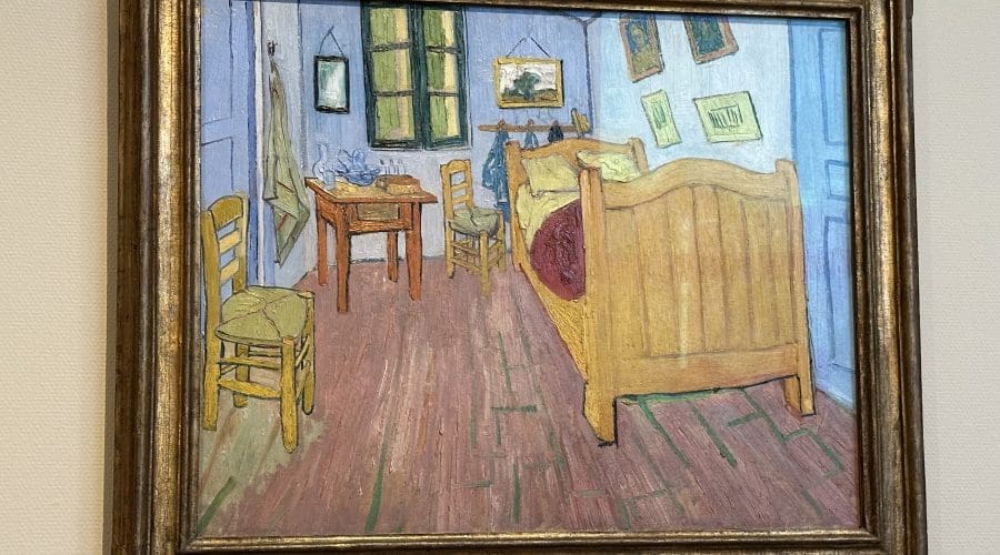 A framed painting depicts a simple bedroom with a wooden bed, chairs, a table, and artwork on the walls, rendered in bold colors and lines. A framed painting depicts a simple bedroom with a wooden bed, chairs, a table, and artwork on the walls, rendered in bold colors and lines.
