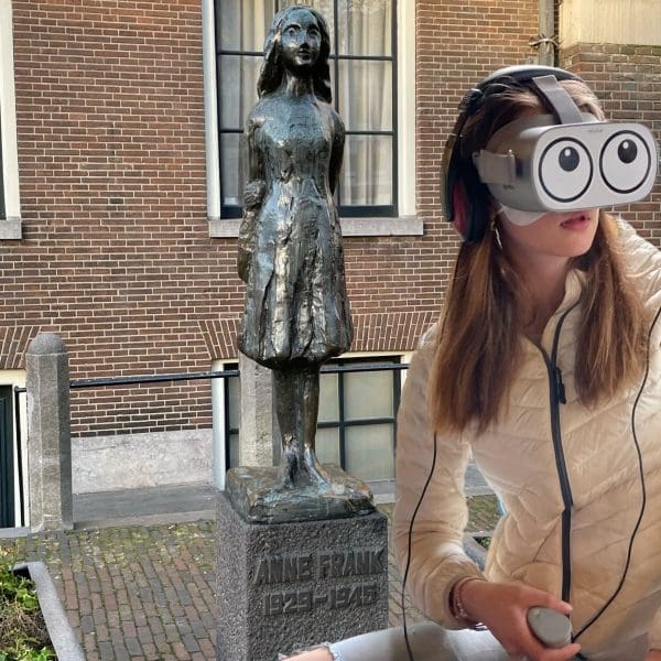 A person wearing a VR headset sits near the Anne Frank statue outside a brick building, immersed in a Virtual Reality Experience that brings the story of the Secret Annex to life amid etched plaques nearby.