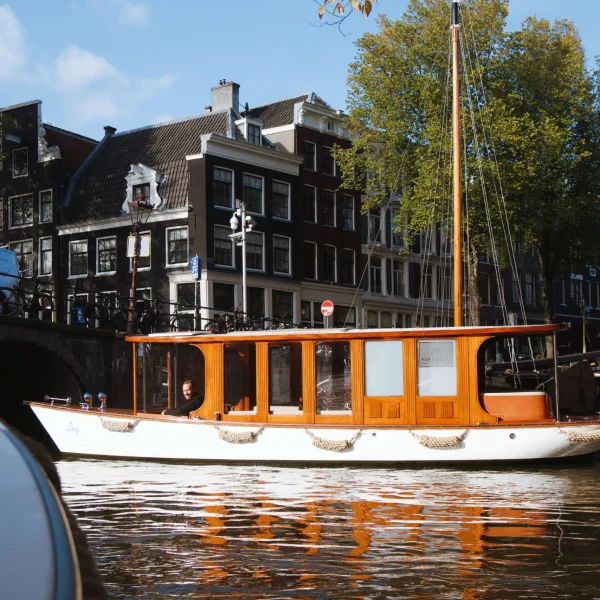 A wooden boat with large windows floats on a canal in front of traditional Dutch buildings and a bridge in Amsterdam, offering the perfect setting for Private Canal Cruises.