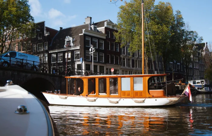 A wooden boat with large windows floats on a canal in front of traditional Dutch buildings and a bridge in Amsterdam, offering the perfect setting for Private Canal Cruises.