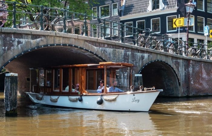 A white canal boat with two people inside passes under a stone bridge in a city, likely on an Amsterdam Canals boat tour, with bikes parked along the railings above.