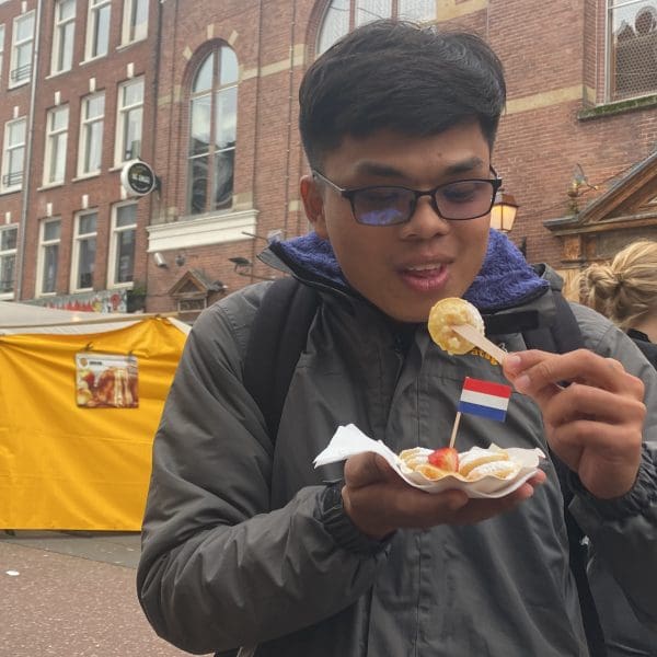 A person holds a small Dutch flag in food while eating street snacks on a city street lined with brick buildings and outdoor market stalls.