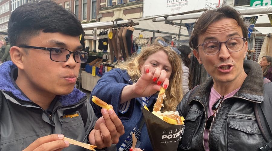 Three people share and eat fries with toppings at an outdoor market, with market stalls and buildings visible in the background. Three people share and eat fries with toppings at an outdoor market, with market stalls and buildings visible in the background.