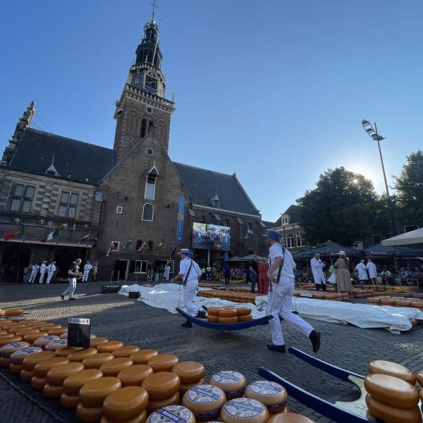 People in white uniforms carry large wheels of cheese in front of a historic building during a cheese market event on a sunny day.